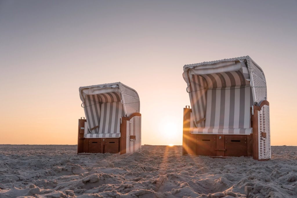 Zwei Strandkörbe an einem Strand an der Nordsee in Nahaufnahme, dahinter geht die Sonne im Meer unter.