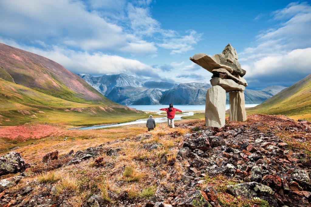 Ein Steindenkmal der Inuit steht im Torngat Nationalpark, dahinter läuft eine ältere Person und ein Kind, das das Steindenkmal imitiert.