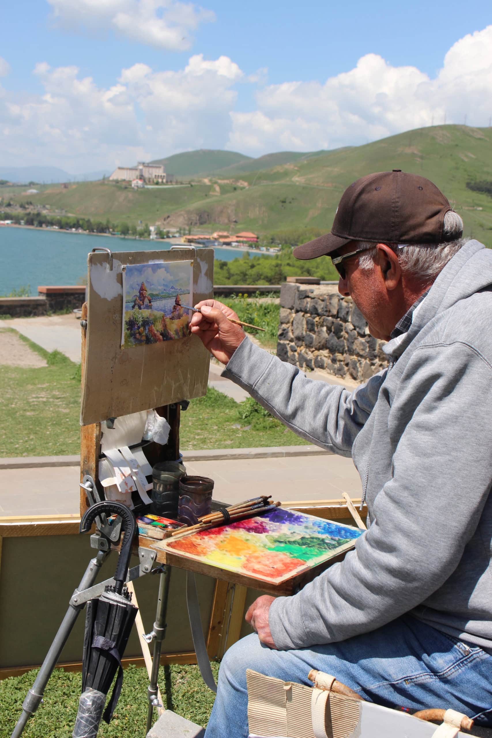 Ein älterer Mann sitzt in Armenien an einer Straße und malt die Szene vor ihm: einen blauen See vor den Bergen des Kaukasus.