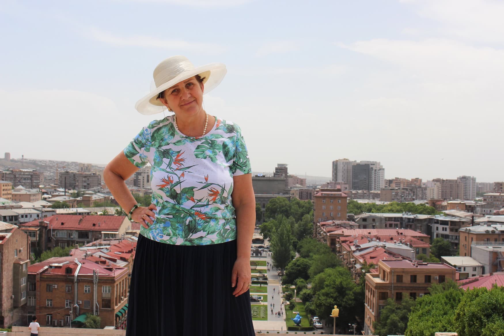 Eine Frau mittleren Alters mit einer Bluse mit Blumenmuster und einem hellen Sonnenhut steht auf einer Dachterasse, im Hintergrund die armenische Hauptstadt Jerewan