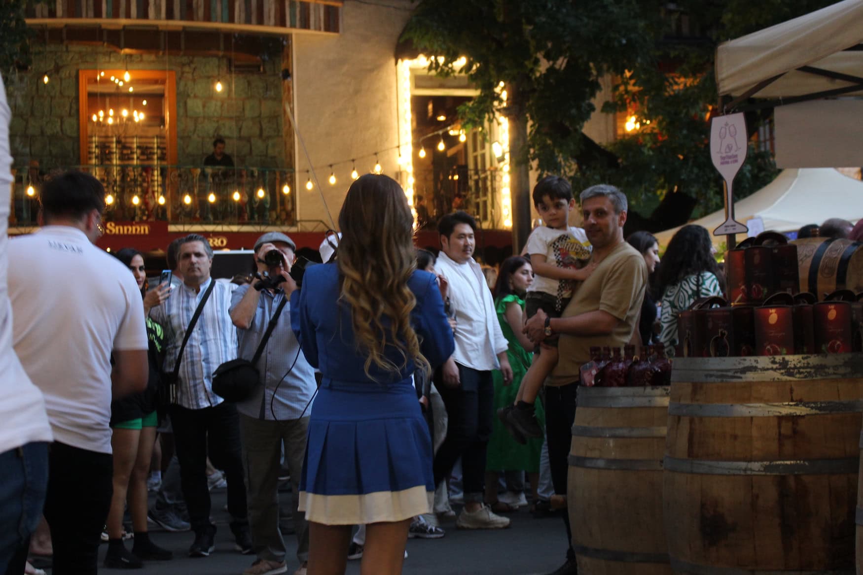 Abendliche Szene auf den Yerevan Wine Days 2025 in Armenien; eine Frau steht mit dem Rücken zur Kamera und wird aus einer Menge im Hintergrund fotografiert. Rechts dient ein aufgestelltes Weinfass einigen Besucher als Stehtisch.