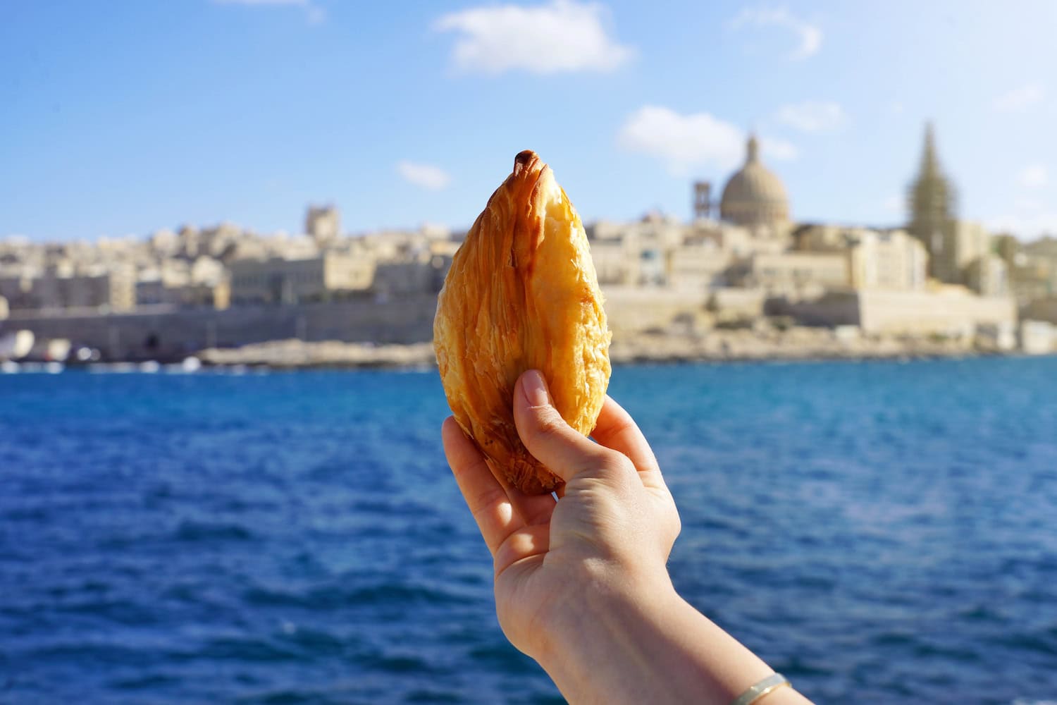 Person hält das Essen Pastizzi in der Hand vor einer Wasserkulisse