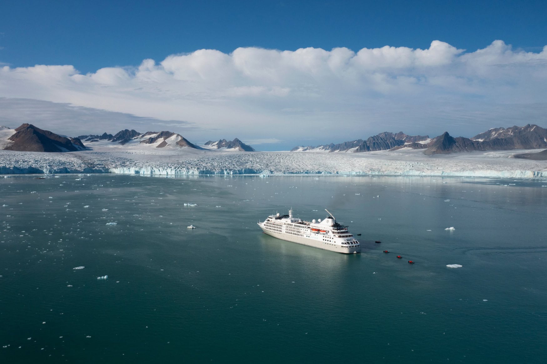 Ein weißes Kreuzfahrtschiff schwimmt im Meer vor einer weiten Eislandschaft, aus der einige felsige Gipfel herausragen.