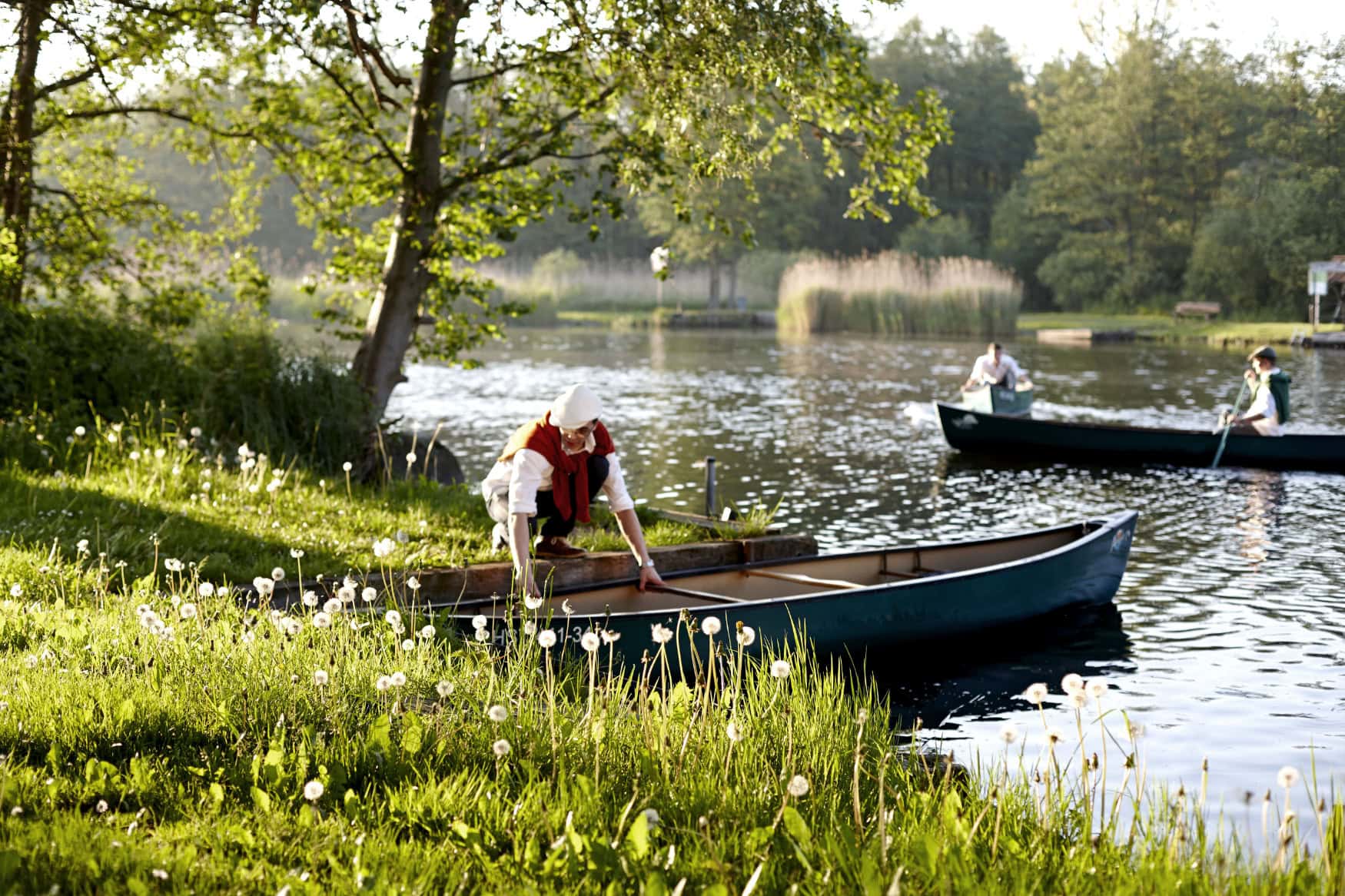Ein Mann in sommerlicher Kleidung steigt am Ufer der Peene in Mecklenburg-Vorpommern in ein Ruderboot. Auf dem Wasser sind bereits zwei weitere Menschen je in einem eigenen Boot unterwegs.
