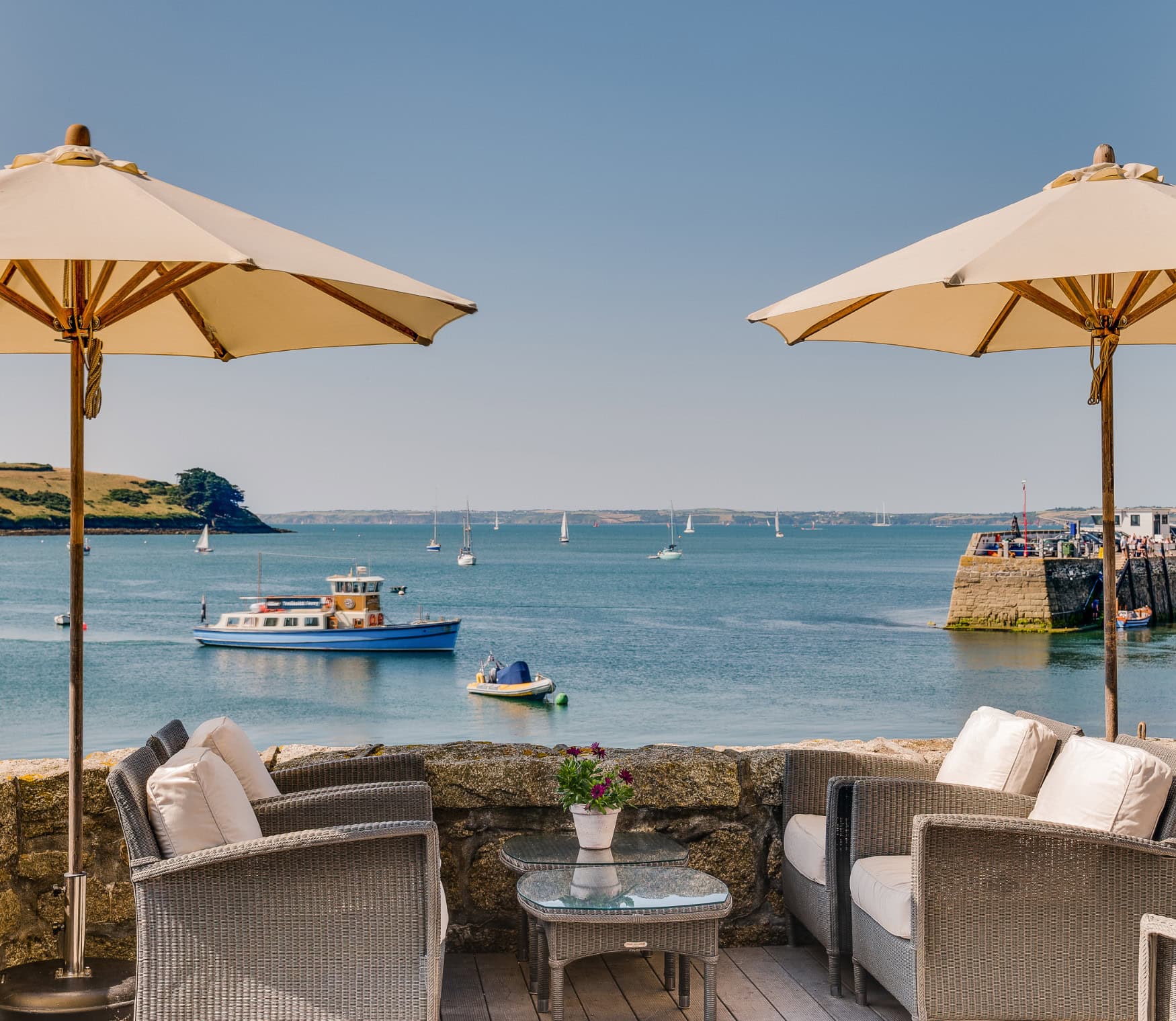 Eine Terrasse mit kleiner Steinmauer, darauf vier graue Korbstühle und zwei Sonnenschirme; dahinter das blaue Meer des Ärmelkanals an der Küste Cornwalls, darauf ein kleines Ausflugsboot und mehrere Segelboote