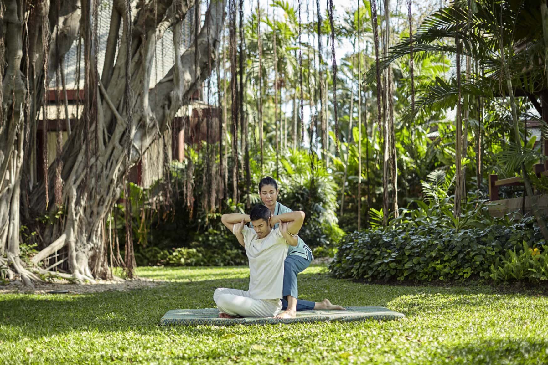 Mitarbeiterin eines Wellness-Resorts in Thailand unterstüzt einen Mann in sommerlicher Kleidung auf einer Übungsmatte im Freien bei Körperübungen; die Szene findet in einem Hotelgarten mit Dschungelatmosphäre statt