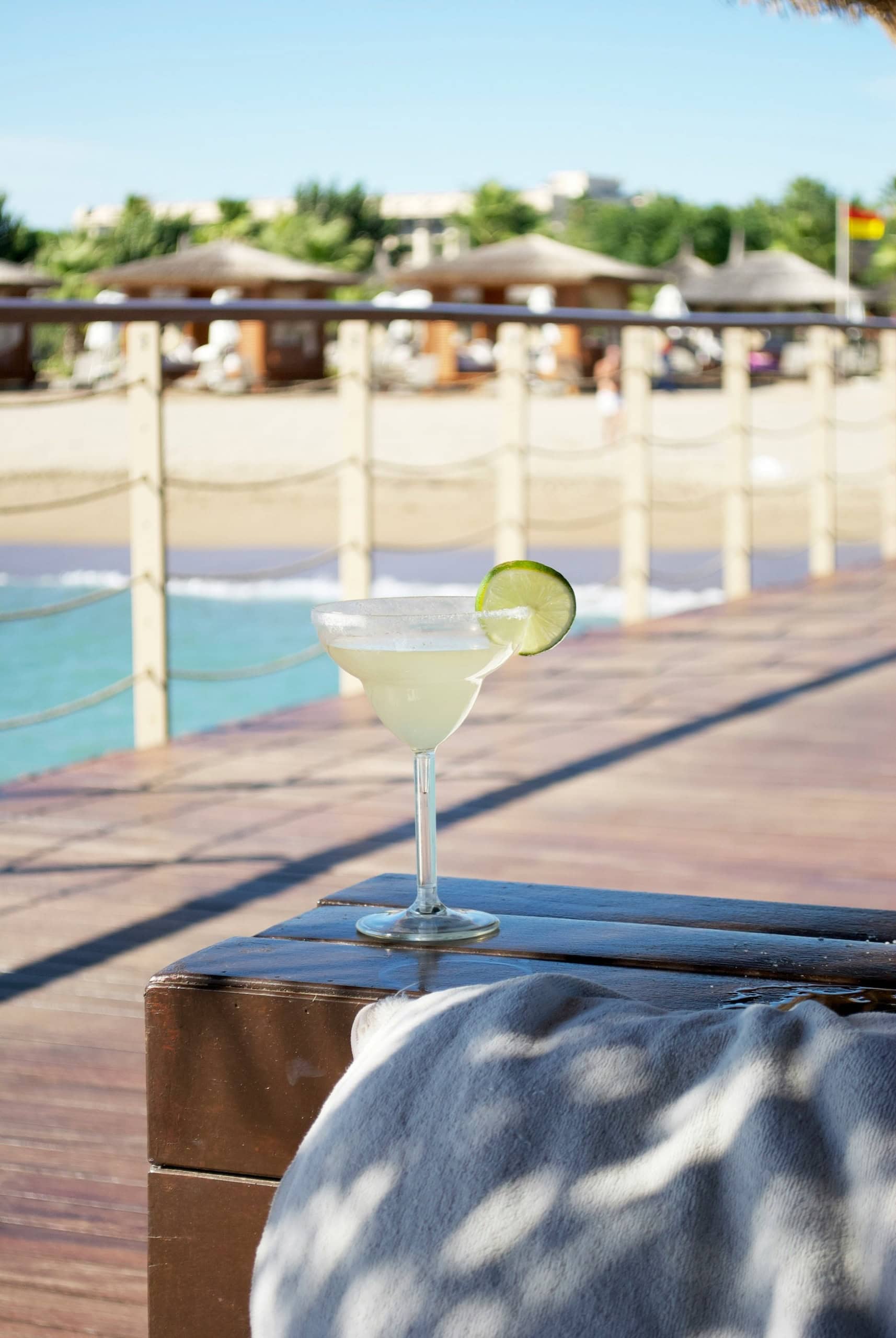 Auf einem Holztisch an einer Strandbar steht ein hohes, rundes Glas mit Margarita, garniert mit einer Limettenscheibe.