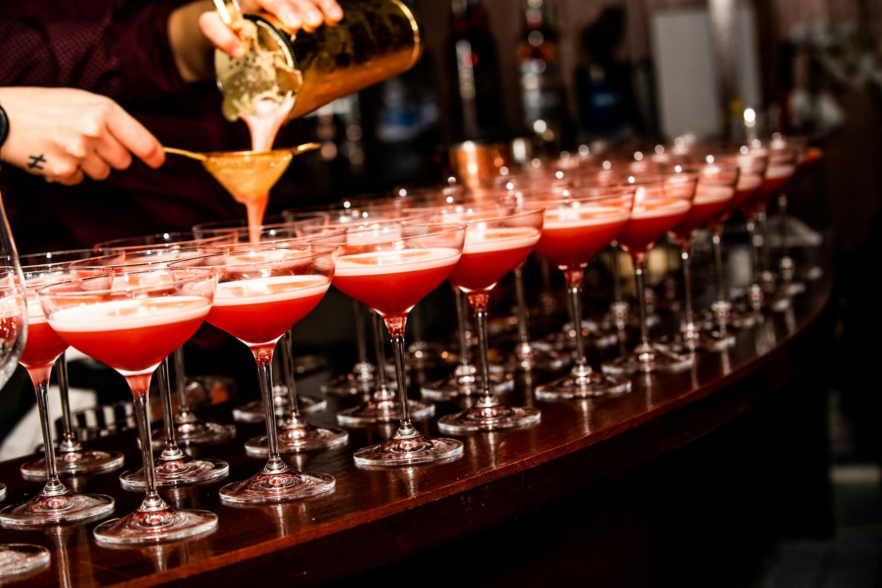 Eine Person bereitet an einer Bar eine Reihe roter Drinks mit Schaum in hohen Stielgläsern vor.