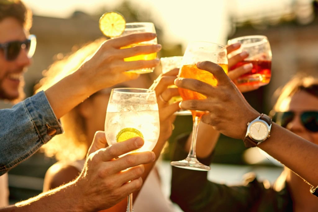 Mehrere Menschen stoßen mit sommerlichen Drinks in der Sonne miteinander an.