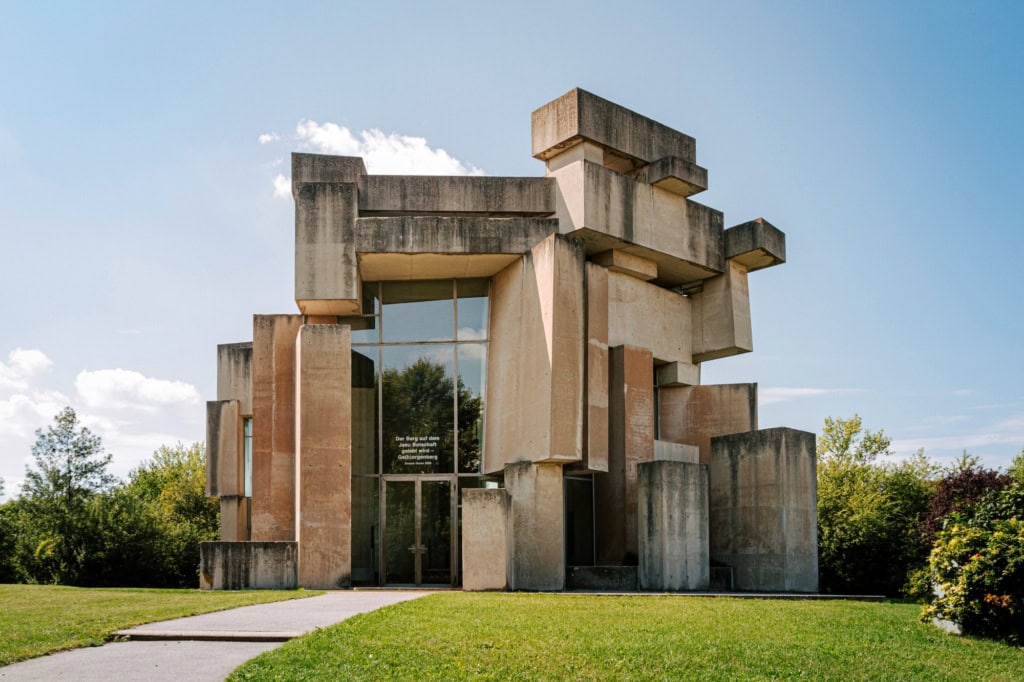 Auf einem grünen Hügel steht eine brutalistische Kirche, die aus zahlreichen großen Betonquadern besteht. Das Einlassportal ist in eine mehrere Meter hohe Glasfront eingelassen.