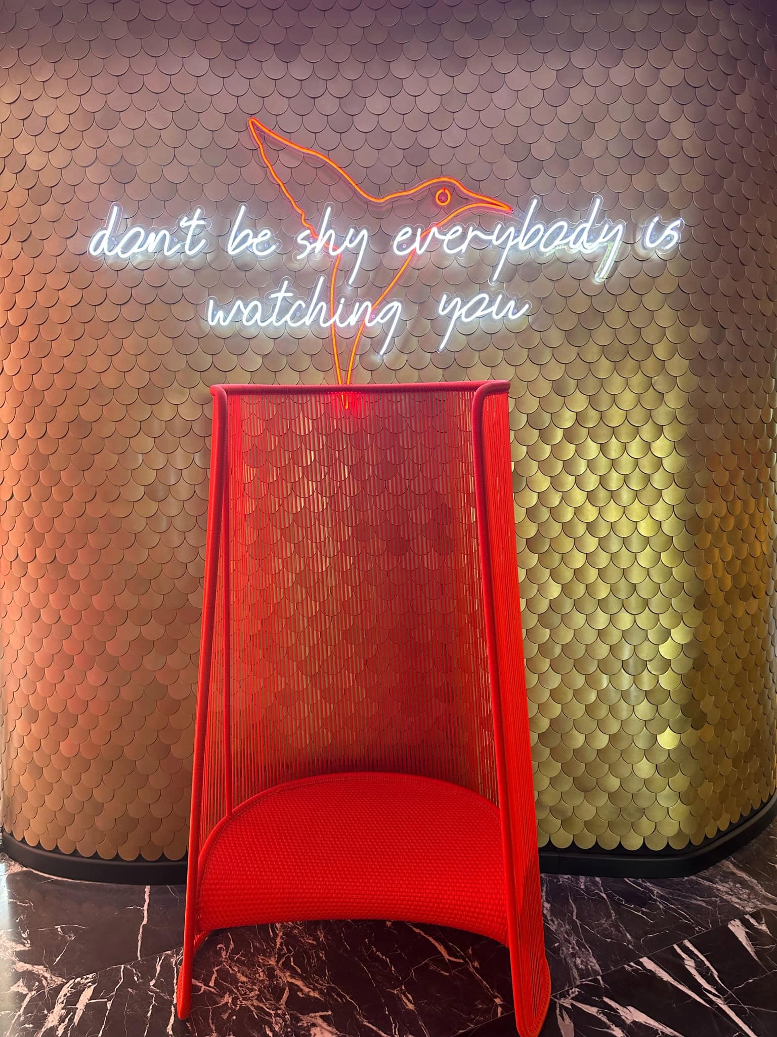 Ein roter Stuhl in der Lobby mit einem Neon Sign „Don't be shy, everybody is watching you"