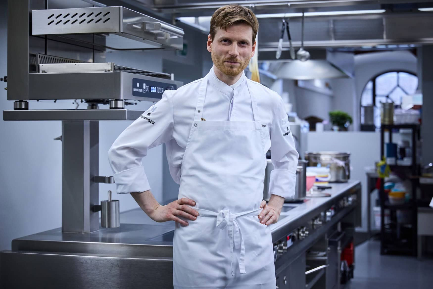 Koch Johannes Nuding steht in einer professionellen Küche und lächelt in die Kamera.