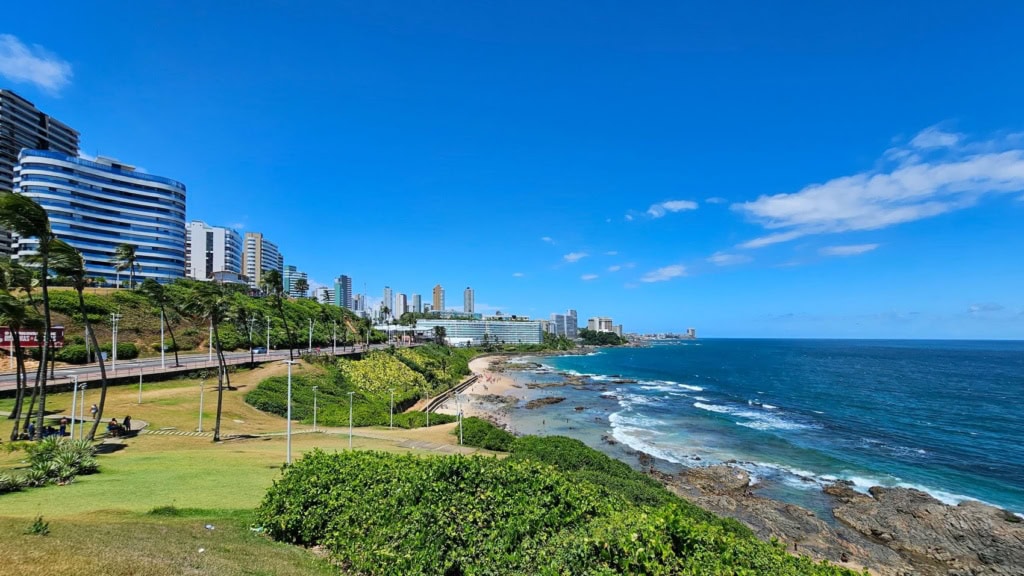 Panoramablick auf die Küste von Salvador da Bahia mit Hochhäusern, Palmen, Grünanlagen und dem Atlantik unter strahlend blauem Himmel.