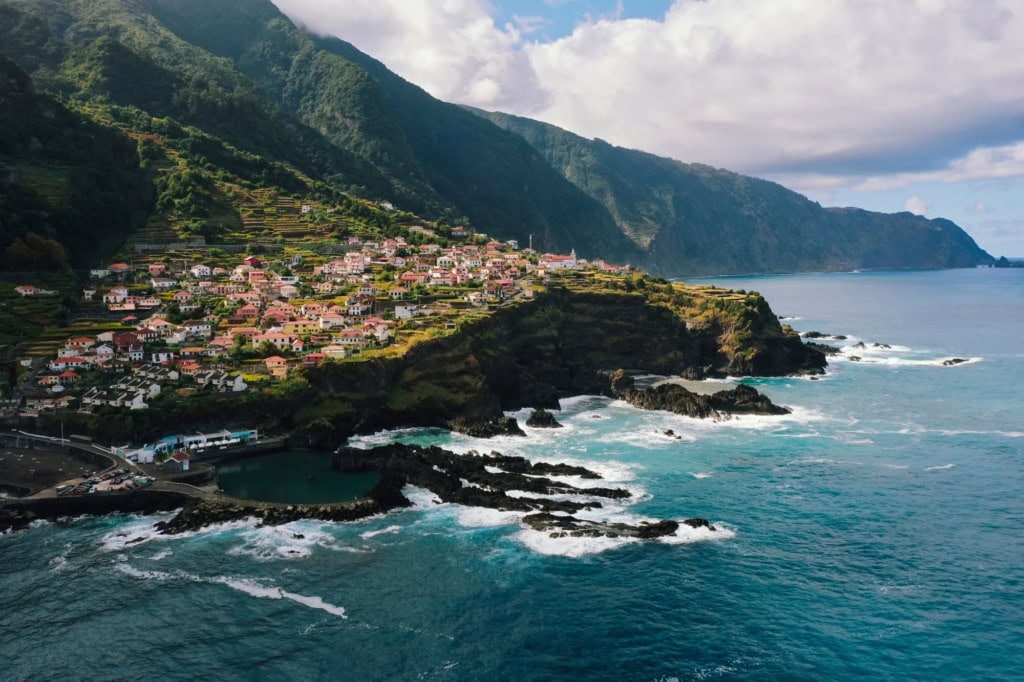 Panoramaaufnahme eines Dorfs an einer Klippe von Madeira; links die Insel mit Gebäuden, rechts der Atlantik.