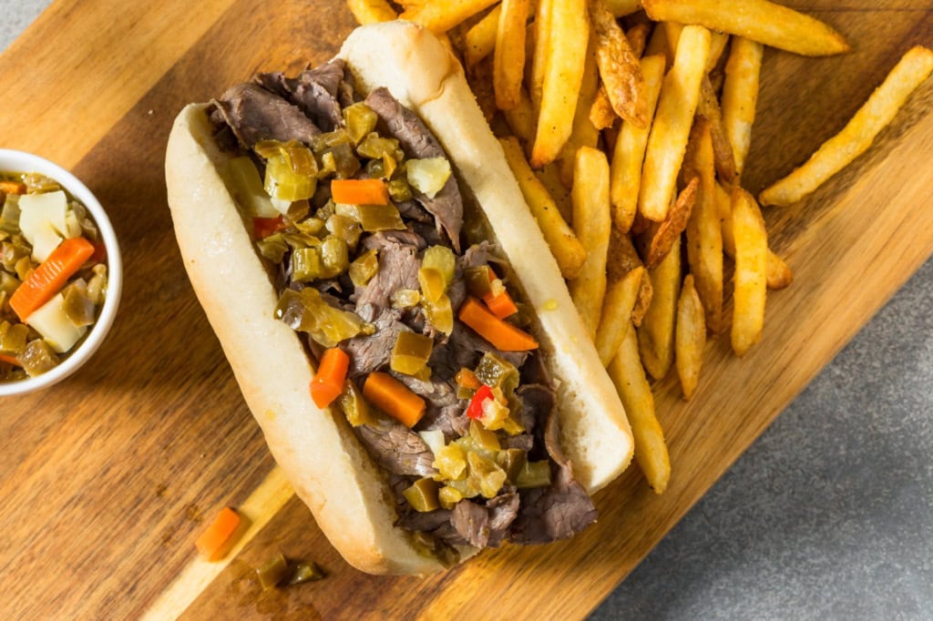 Auf einem Holzbrett liegt ein Italian Beef Sandwich Chicago Style, daneben eine Portion Pommes.