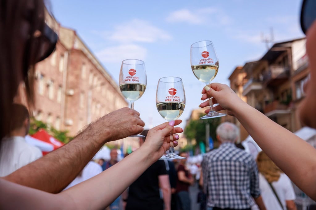 Drei Hände stoßen mit drei Weingläsern, jeweils mit der Aufschrift "Yerevan Wine Days" an; unscharf dahinter eine Szene auf einem Straßenfest in Armenien.