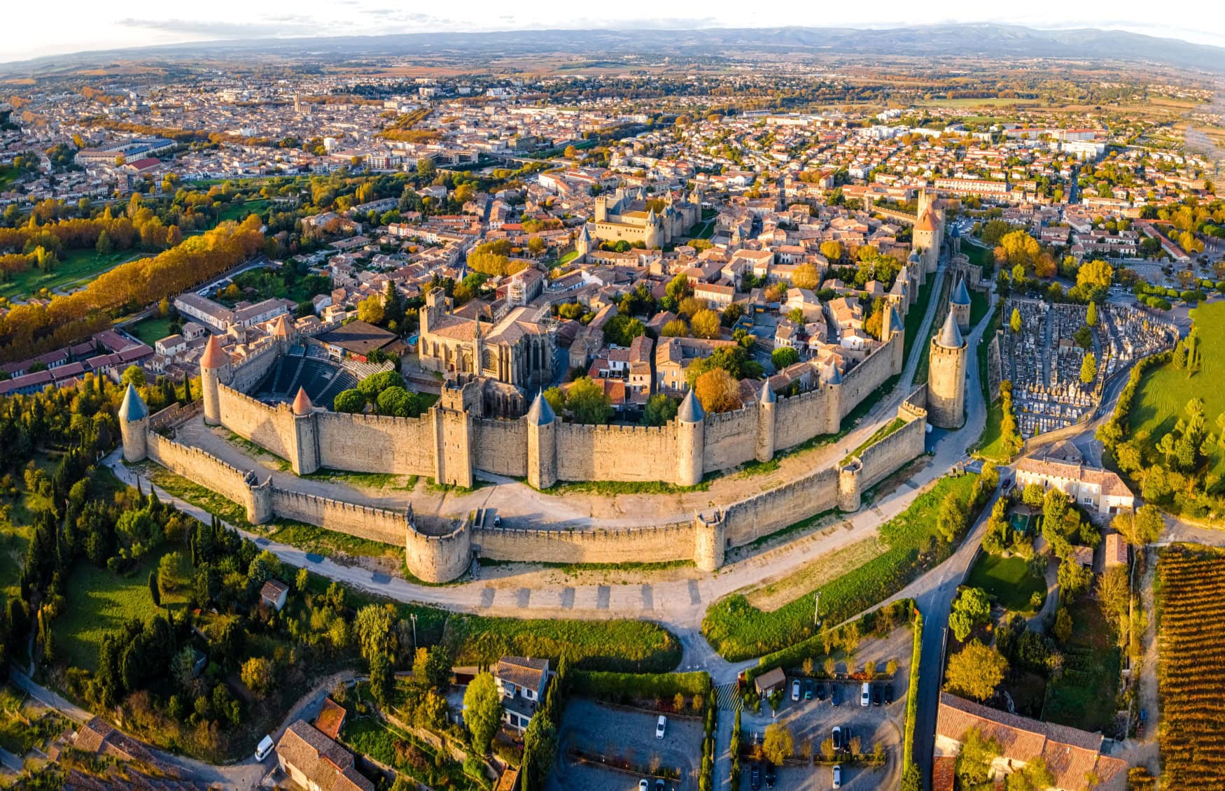 Luftaufnahme der Festungsstadt Carcassonne in Südfrankreich; die breite Doppelmauer zeigt zum Betrachter, in der Stadt selbst sind die Kathedrale und die alte Burg gut im Sonnenuntergang zu sehen.
