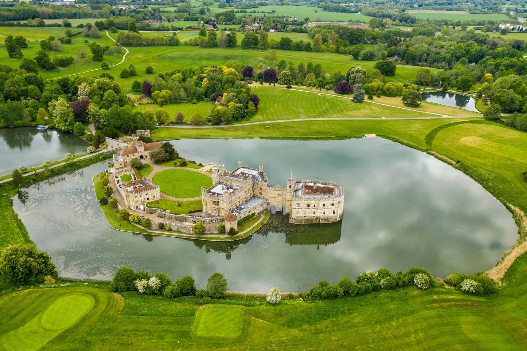 Inmitten eines kleinen Sees in einer wohlgepflegten Parklandschaft liegt eine längliche Burg mit mehreren Wohnbauten auf einer Insel. Ein Teil der Burg ragt sogar direkt aus dem Wasser. Aufgenommen mit einer Drohne.