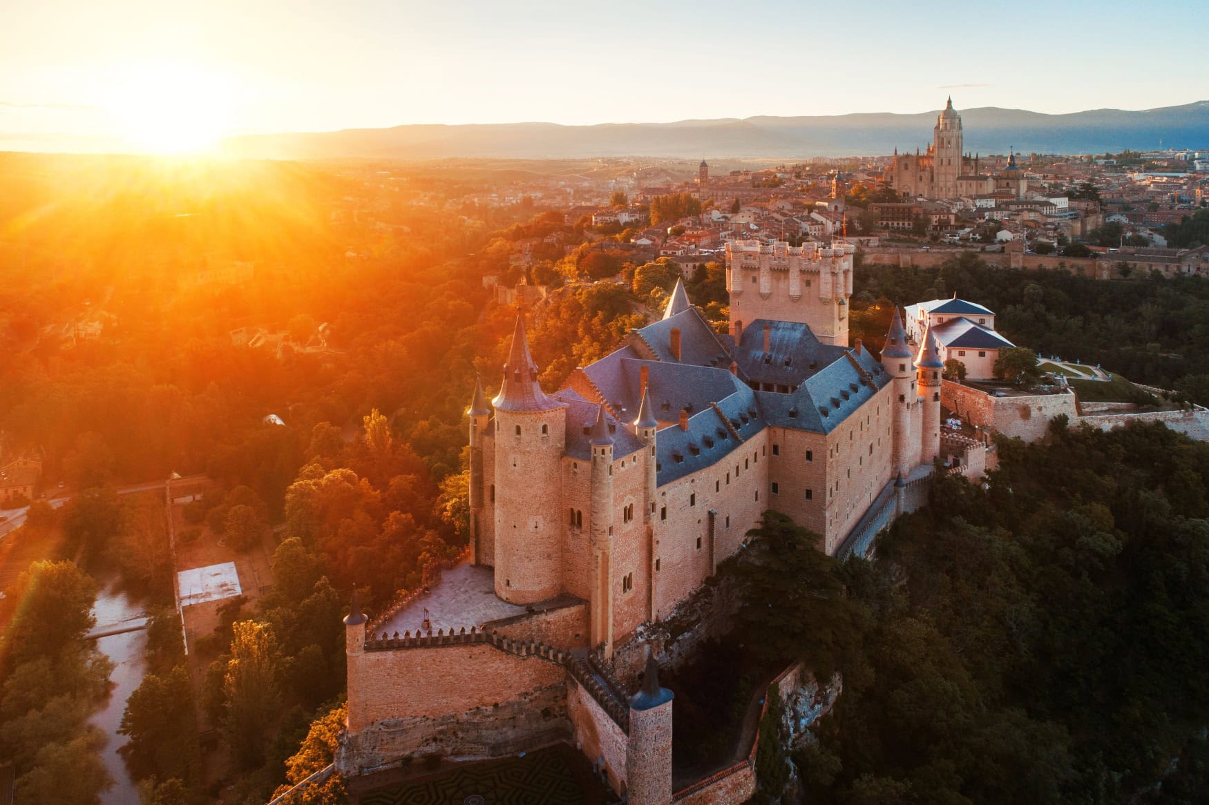 Die mächtige Burg Alcazar von Segovia thront auf einem Felsvorsprung. Im Hintergrund ist die Stadt sichtbar, links am Horizont geht die Sonne unter.