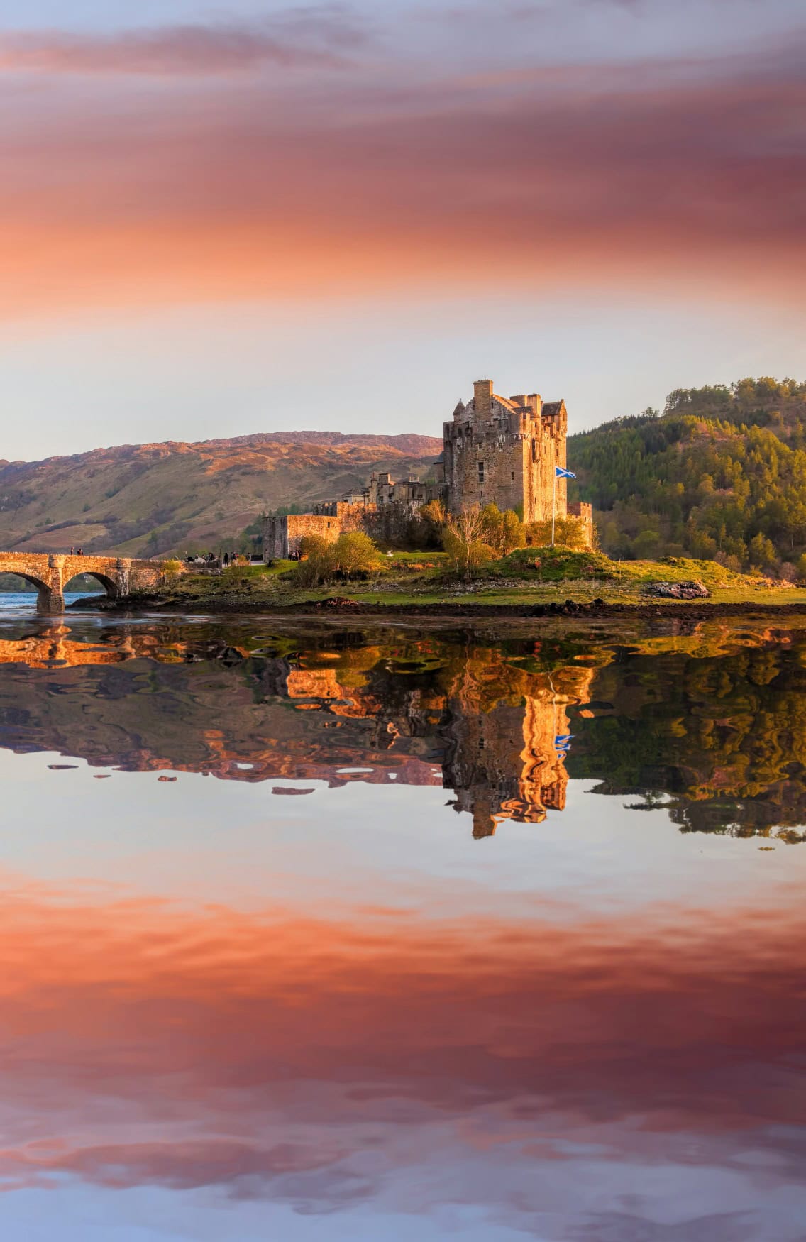 In einem schottischen See liegt eine kleine Insel, darauf ein altes, hohes Schloss. Schloss und See liegen im roten Licht der Abendsonne, auf der stillen Wasseroberfläche spiegelt sich das Bild.