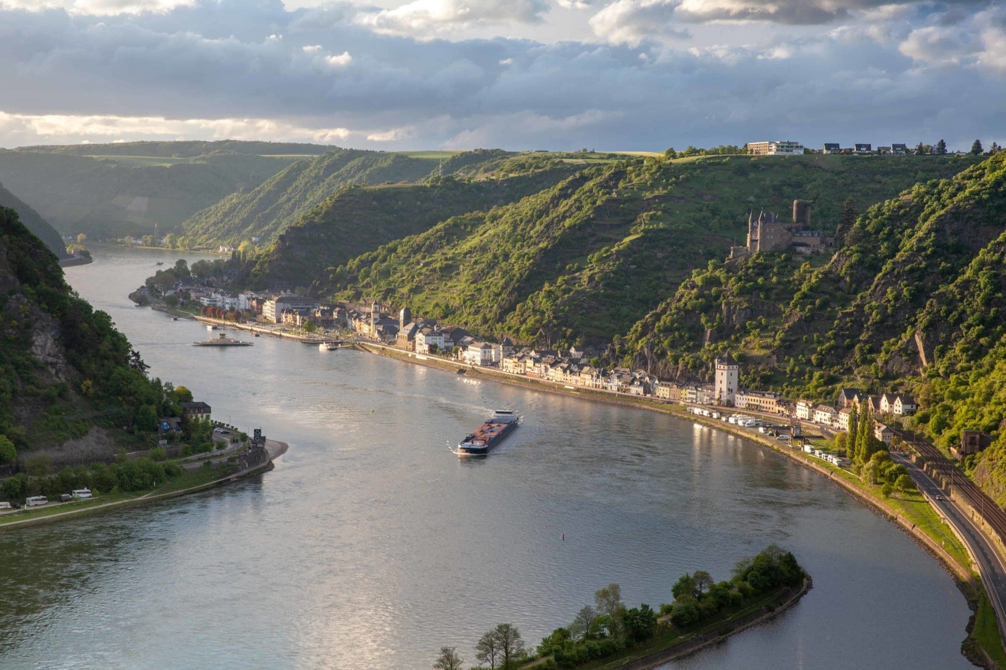 Von einer Anhöhe aus zieht sich der Mittelrhein in einer S-Schleife durch das Rheintal. Auf dem Rhein ist ein Frachtschiff unterwegs. Am Hang zur Rechten thront über einer kleinen Stadt eine gut erhaltene Burg. Alles ist sommerlich grün.