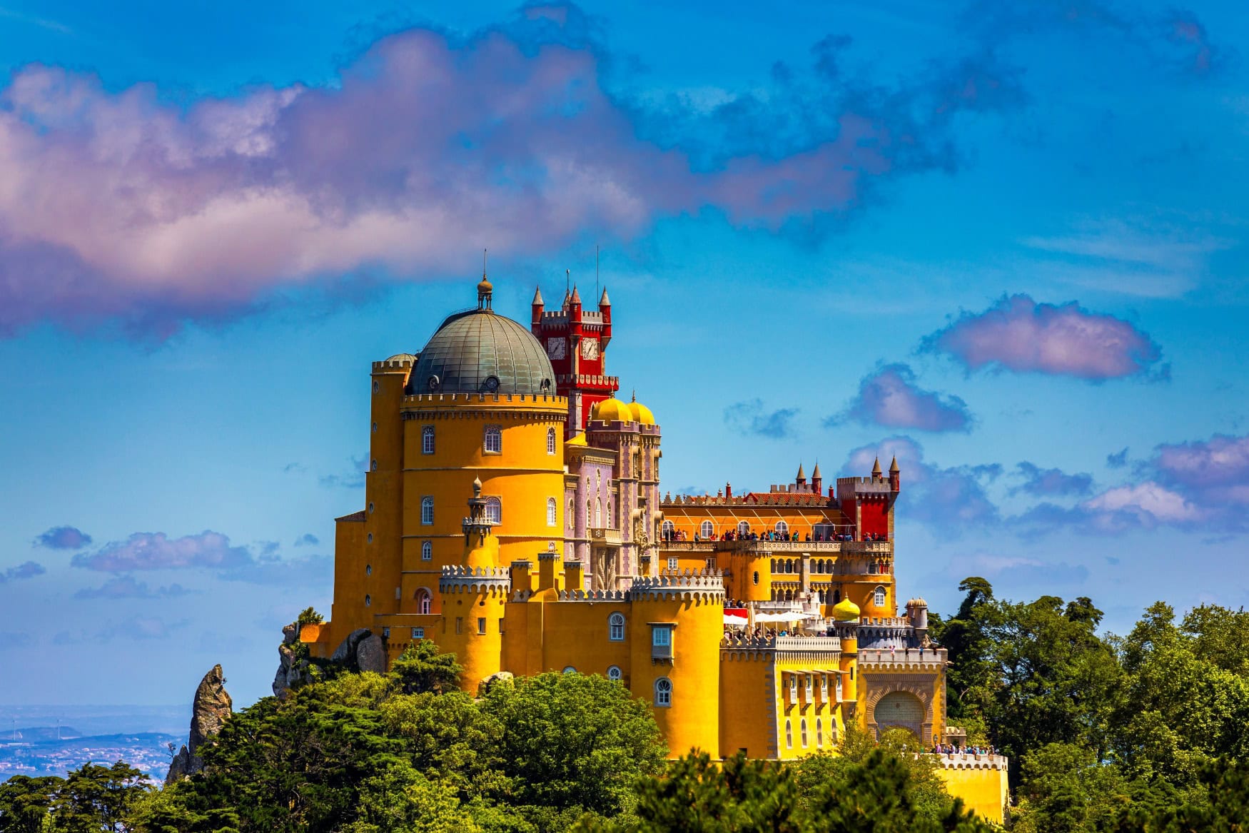 Ein historistisches, gelbes Märchenschloss thront auf einem bewaldeten Felsvorsprung unter blauem Himmel.