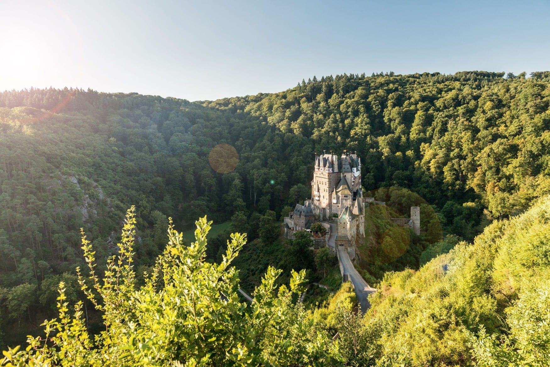 Von einer bewaldeten Anhöhe geht der Blick in ein grünes Tal, in dessen Mitte sich Burg Eltz erhebt. Von links scheint die Sonne ins Bild und erzeugt einen Linseneffekt.