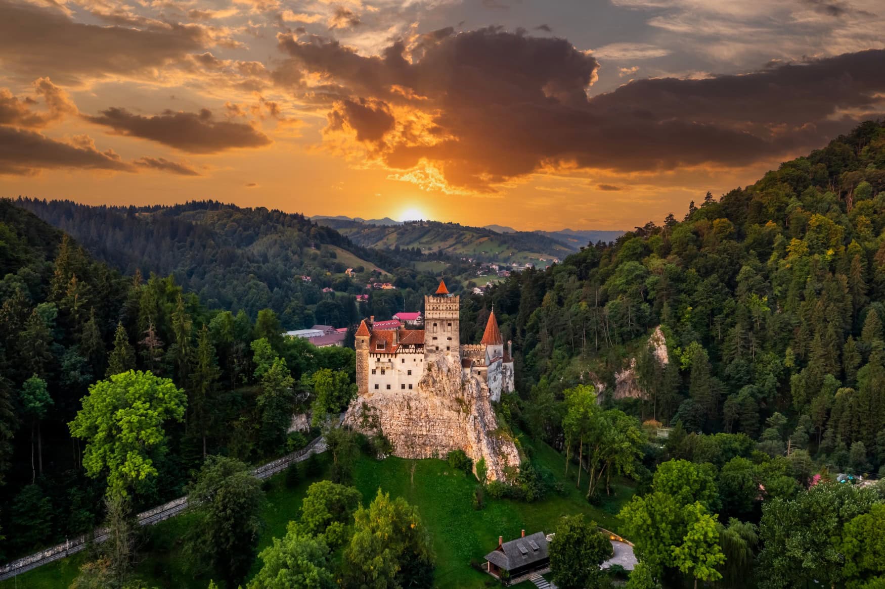 Drohnenaufnahme einer Spornburg in Rumänien, dem Schloss Bran. Hinter der Burg liegt ein verwinkeltes Tal, über einem grünen Hügel am Horizont geht die Sonne unter.