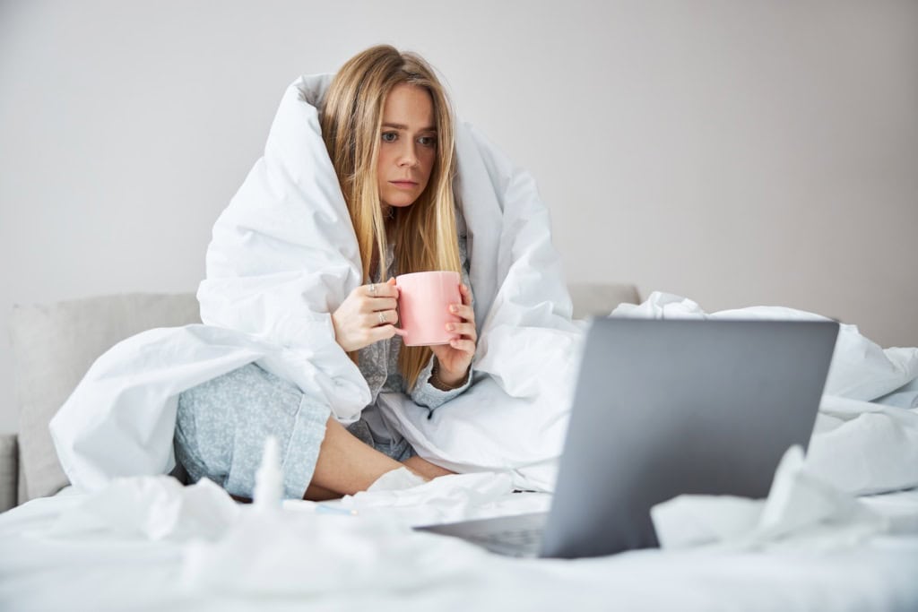 Eine kranke Frau sitzt im Bett vor ihrem Laptop.