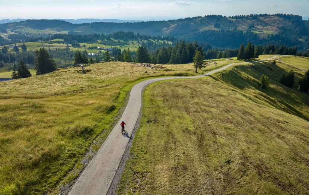 Drohnenaufnahme einer Person, die auf einem langezogen Radweg entlang einer grüner Hügel im Allgäu unterwegs ist.