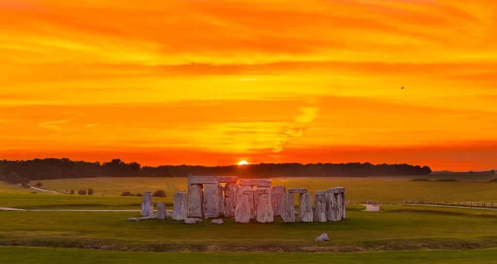 Luftaufnahme von Stonehenge in England; im Hintergrund geht über einem Waldstück rot-gelb die Sonne unter.