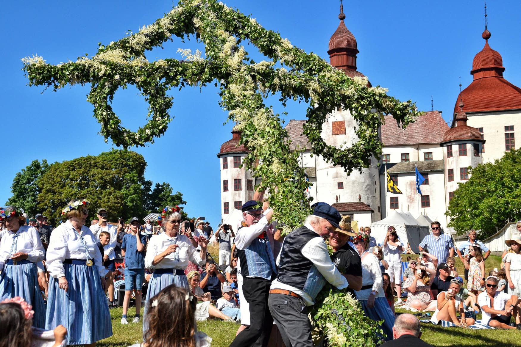 Mehrere Männer in traditioneller Kleidung stellen auf einer großen Mittsommer-Feier in Schweden einen grün geschmückten Pfahl auf.
