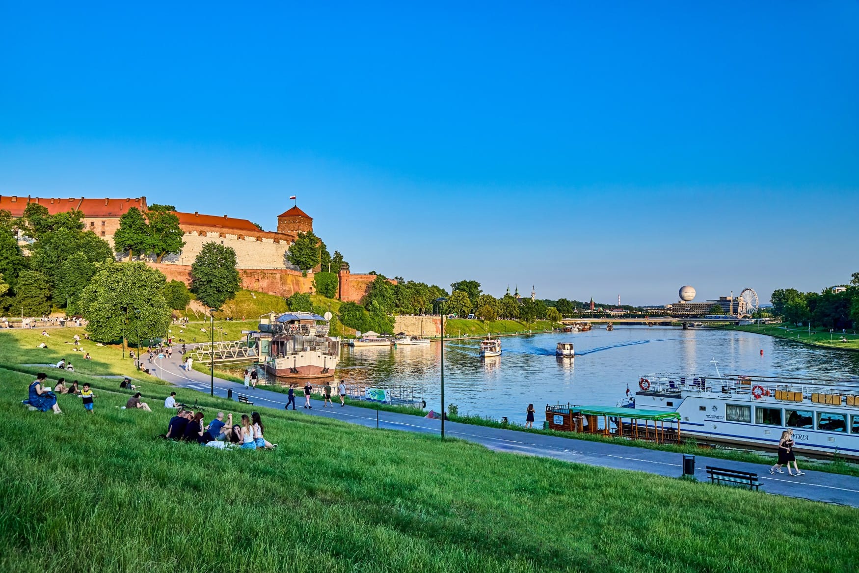Auf einem begrünten Flussufer sitzen mehrere Menschengruppen inder Sonne; links im Hintergrund erhebt sich der Wawel, auf dem Fluss Weichsel fahren mehrere Schiffe