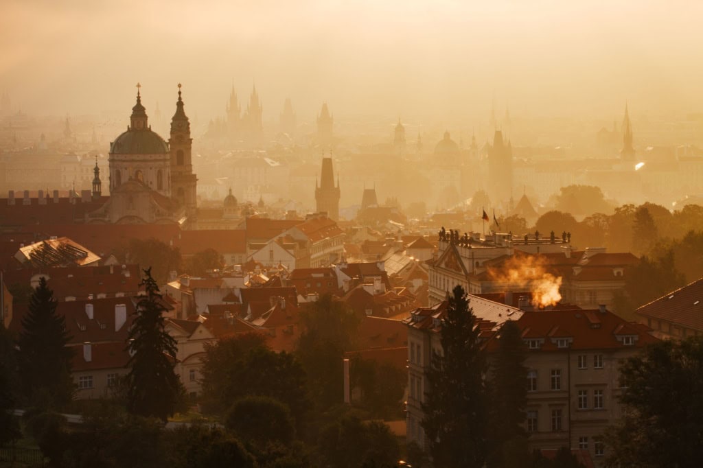 Ein Meer aus Dächern. Am linken Rand ragt prominent die messingfarbene Kuppel einer Kirche empor, rechts der quadratische Dachfirst eines Palais, weit im Hintergrund die Doppeltürme einer Kathedrale. Das gesamte Bild ist von der aufgehenden Sonne in ein rot-goldenes Licht getaucht und sehr diesig, der Hintergrund ist kaum noch zu erkennen