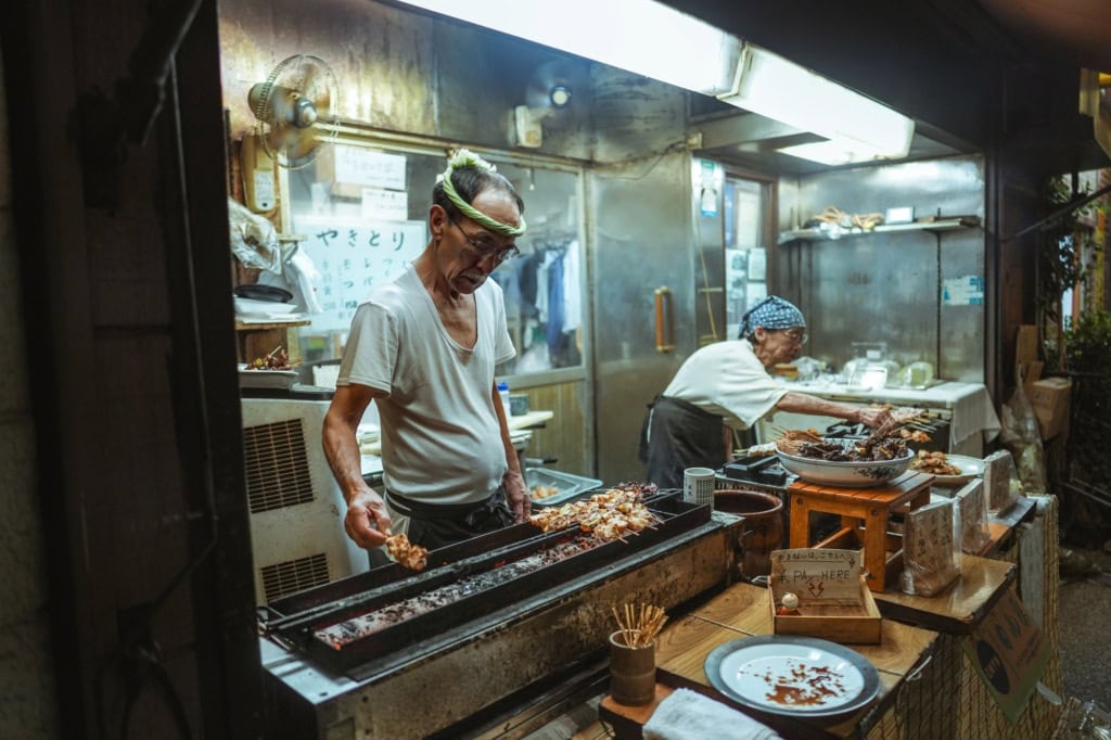 Ein älterer Mann und eine ältere Frau betreiben gemeinsam einen kleinen Imbissstand in Tokio und verkaufen Yakitori-Spieße.
