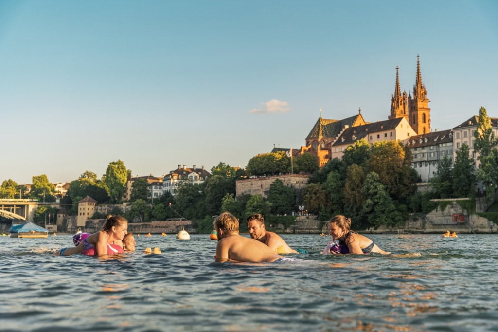 Eine Gruppe junger Menschen schwimmt an einem Sommertag im Rhein in Basel; im Hintergrund ragt der Dom aus den Dächern der Stadt hervor.