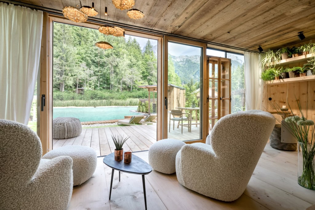 Zwei flauschige Polstersessel mit Hockern stehen in einem Raum, dessen Decken und Boden aus hellen Dielen bestehen. Sie sind auf eine bodentiefe Glaswand ausgerichtet, hinter der eine Holzveranda, ein natürlicher Pool und dann ein Waldstück mit einigen Bergen dahinter sichtbar werden.