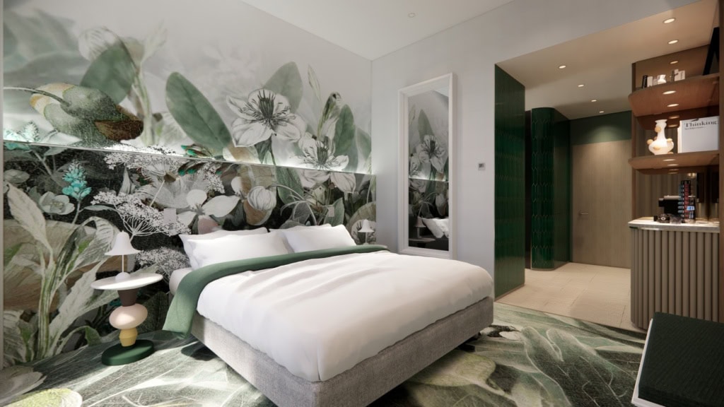 Ein Hotelzimmer; mittig vor einer Wand mit großflächigem Blumenmusterdruck mit Grün- und Weißtönen steht ein breites Doppelbett mit weißen Laken. Auf dem Boden ein großer, dunkelgrüner Teppich. Rechts im Bild schließt sich der Flurbereich an, ganz rechts eine kleine holzverkleidete Bar mit Kaffeemaschine.