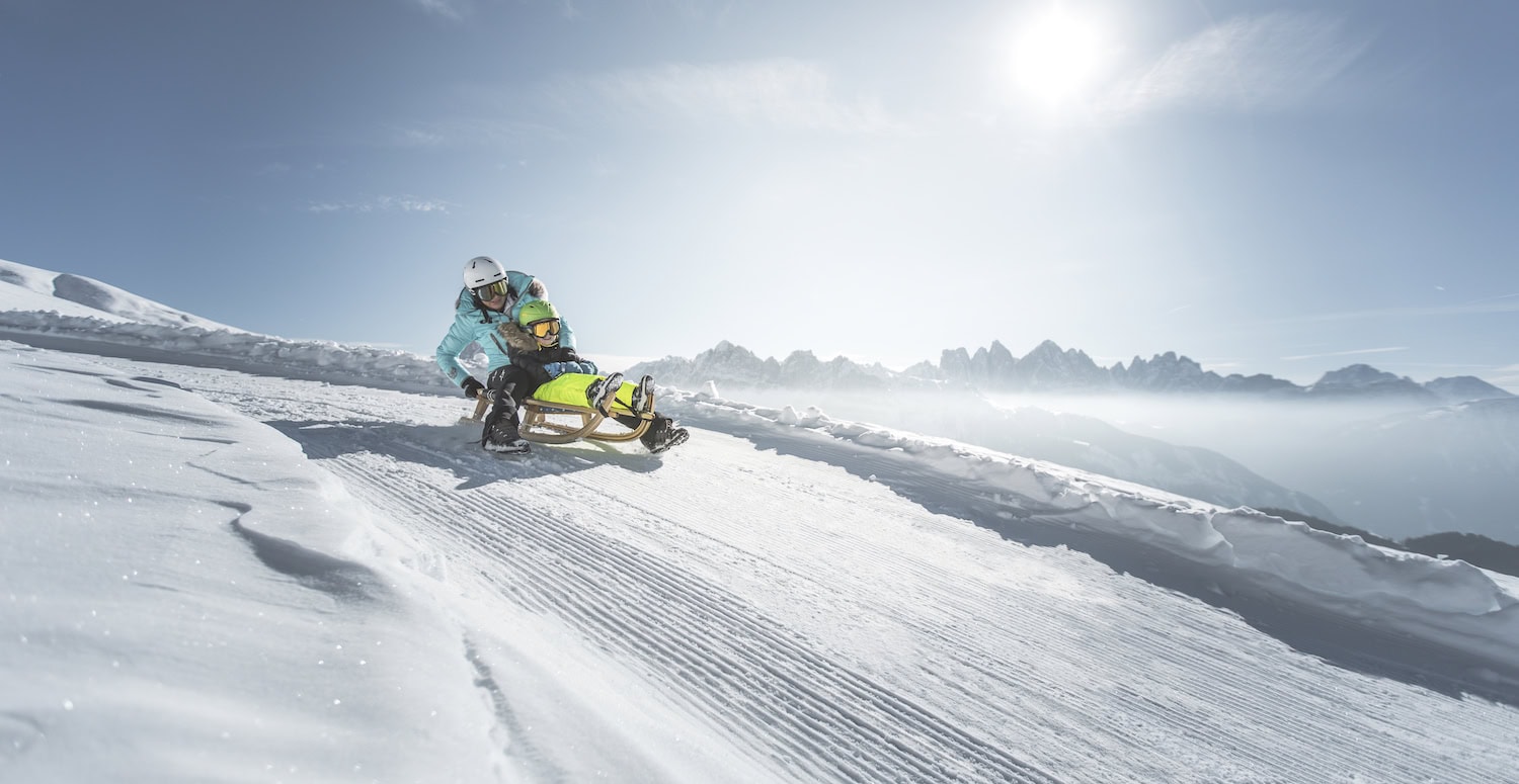Mann und Kind auf Schlitten, rodelnd über eine sonnige Schneepiste, im HIntergrund Berge