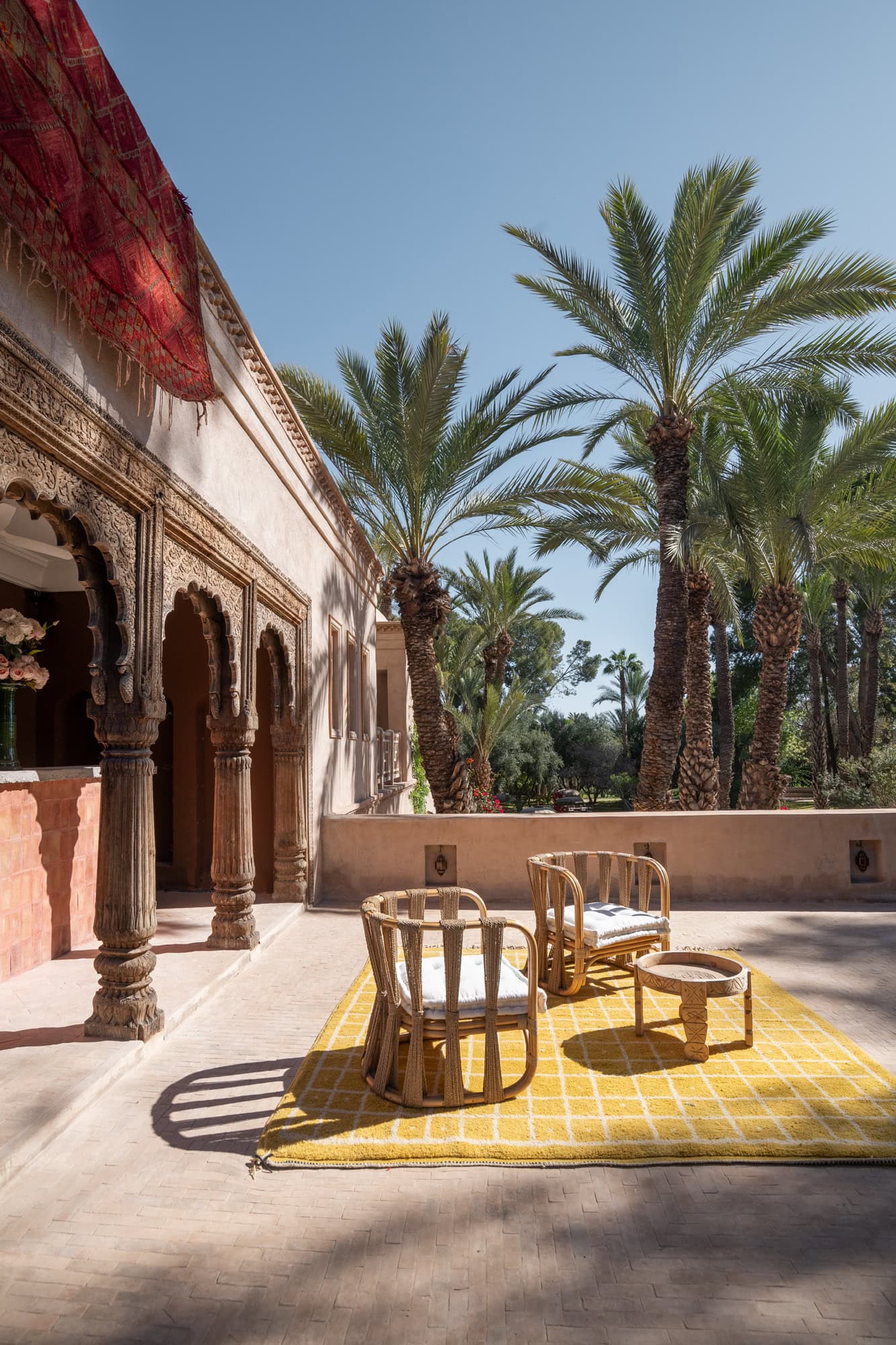 Terrasse in der Sonne mit Palmen und blauem Himmel, zwei Stühlen und einem Tisch auf einem gelben Teppich in Marrakesch