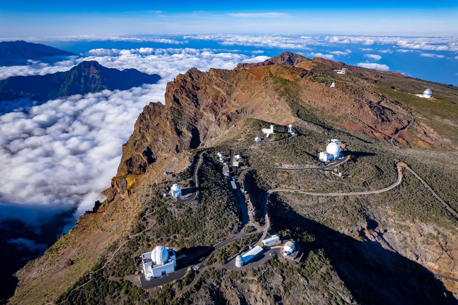 La Palma Observatorium Roque de Los Muchachos - Luftaufnahme auf den Berg mit den darauf verteilten Gebäuden des Observatoriums, im Hintergrund tiefhängende Wolken und ein blauer Himmel