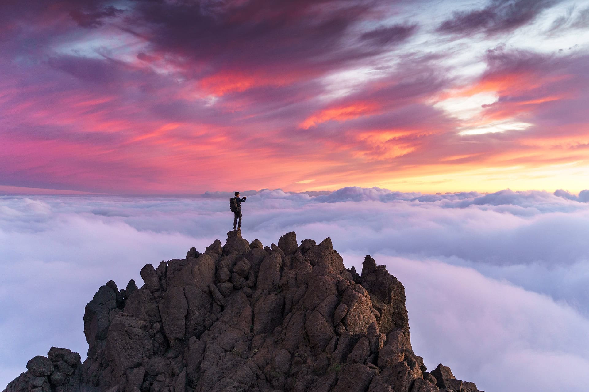Mann auf einsamen Gipfel vor einem dramatischen Sonnenuntergangshimmel, der das Panorama mit dem darunterliegenden Wolkenmeer fotografiert.
