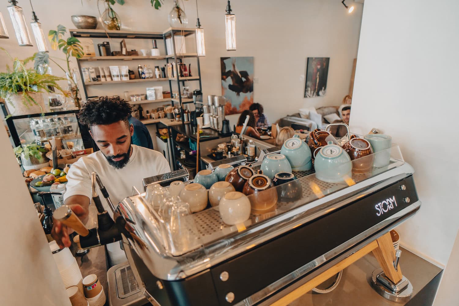 Barista vor großer Kaffeemaschine, im Hintergrund Gäste des Cafes