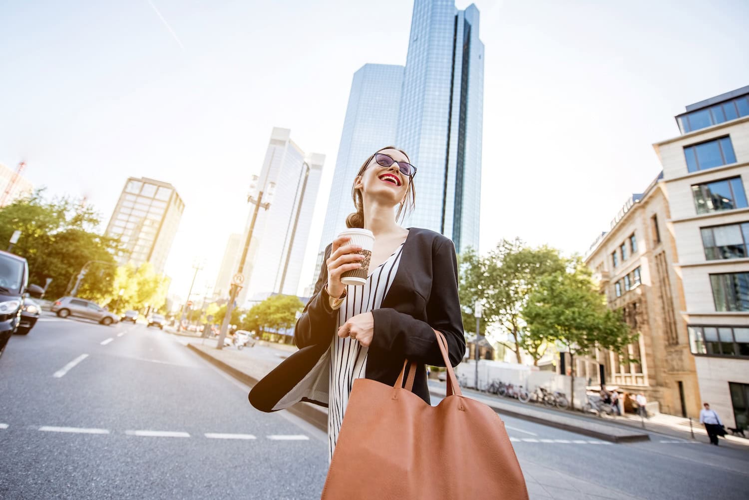 Lachende Frau mit Kaffee in der Hand auf einer Straße in Frankfurt mit einem Hochhaus im Hintergrund
