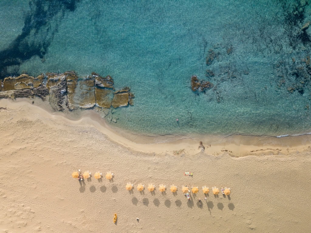 Luftaufnahme eines hellen Sandstrands mit türkisfarbenem, klarem Wasser und Felsen im Meer. Mehrere Sonnenschirme stehen in einer Reihe am Strand, vereinzelt liegen Menschen im Sand oder schwimmen im Wasser