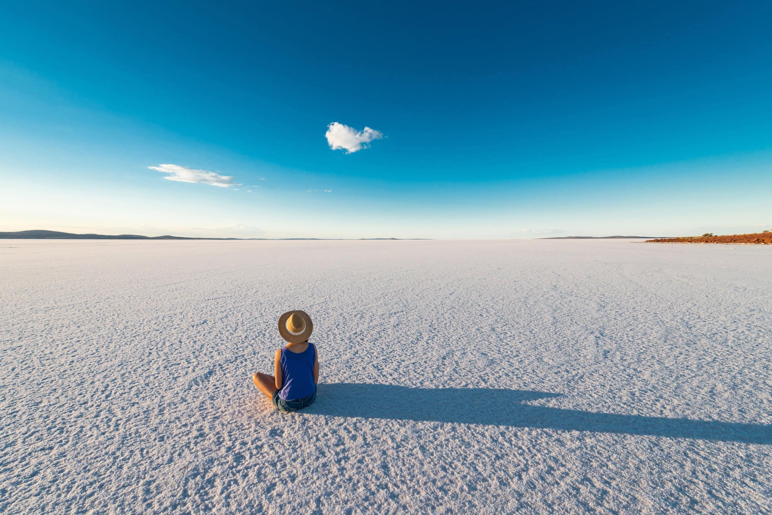 Der Lake Gairdner in Südaustralien; ein Salzsee, der bis zum Horizont reicht. Im Vordergrund sitzt eine Frau mit Sonnenhut im Schneidersitz auf dem Boden