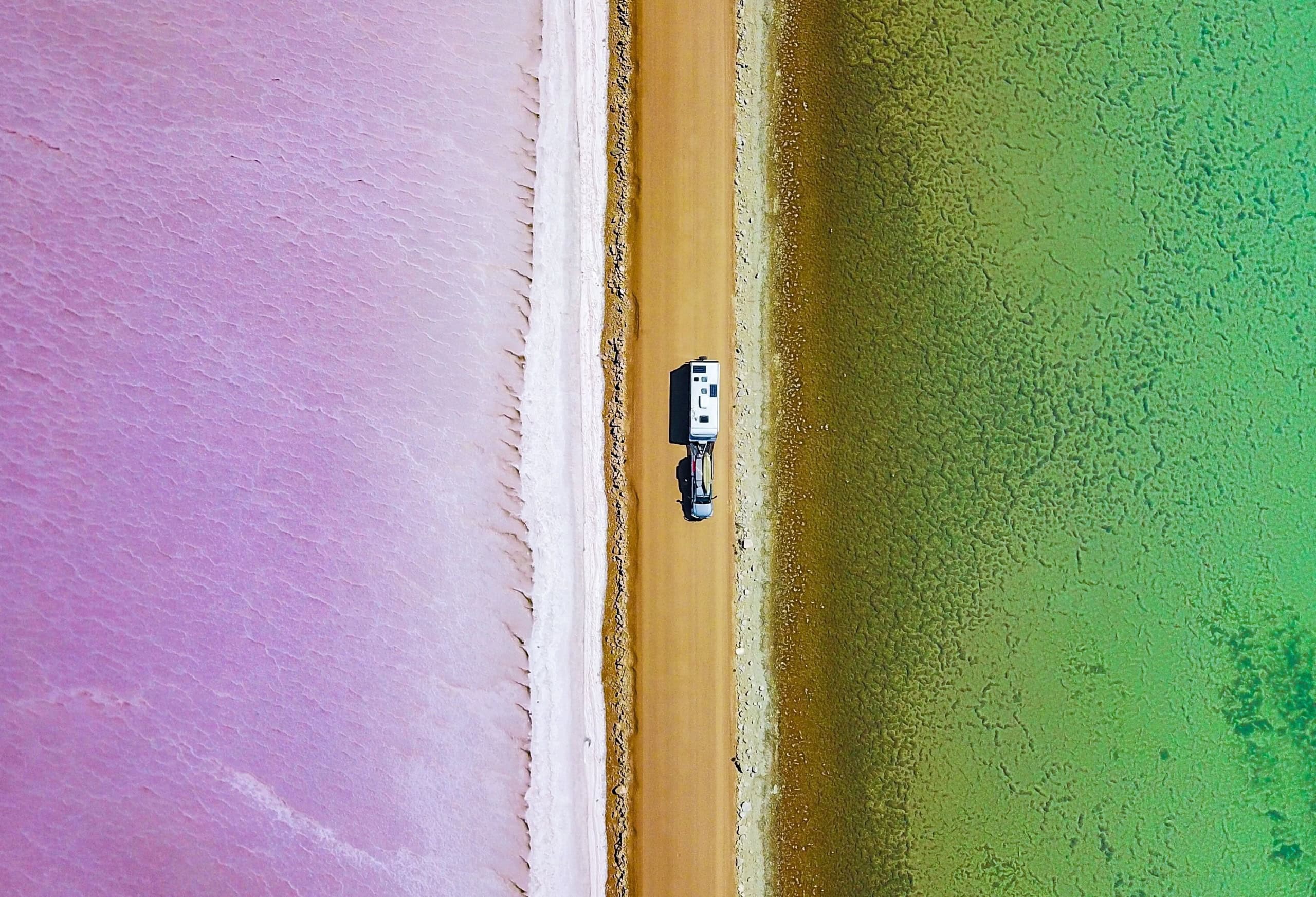Der Lake MacDonnell ist ein Must-see im Südaustralien-Urlaub; Luftaufnahme eines Autos mit Wohnwagen, das auf einer braunen Piste genau von oben nach unten zwischen zwei Seen fährt, der links ganz in violett, der rechte ganz grün