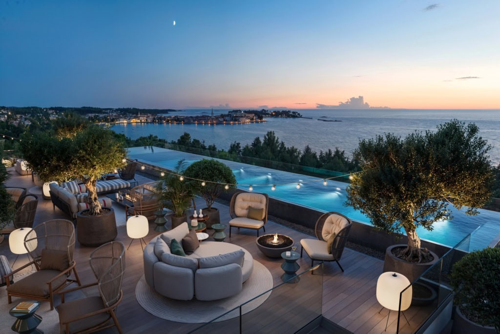 Blick auf die Rooftop-Bar eines Strandhotels am Abend. Zur Linken die Terrasse mit mehreren Sitzgruppen, Polster- und Bastmöbel gemischt. Rechts dahinter ein langezogener Infinitypool. Am Horizont ist die Stadt Porec zu sehen, dahinter das blaue Mittelmeer.