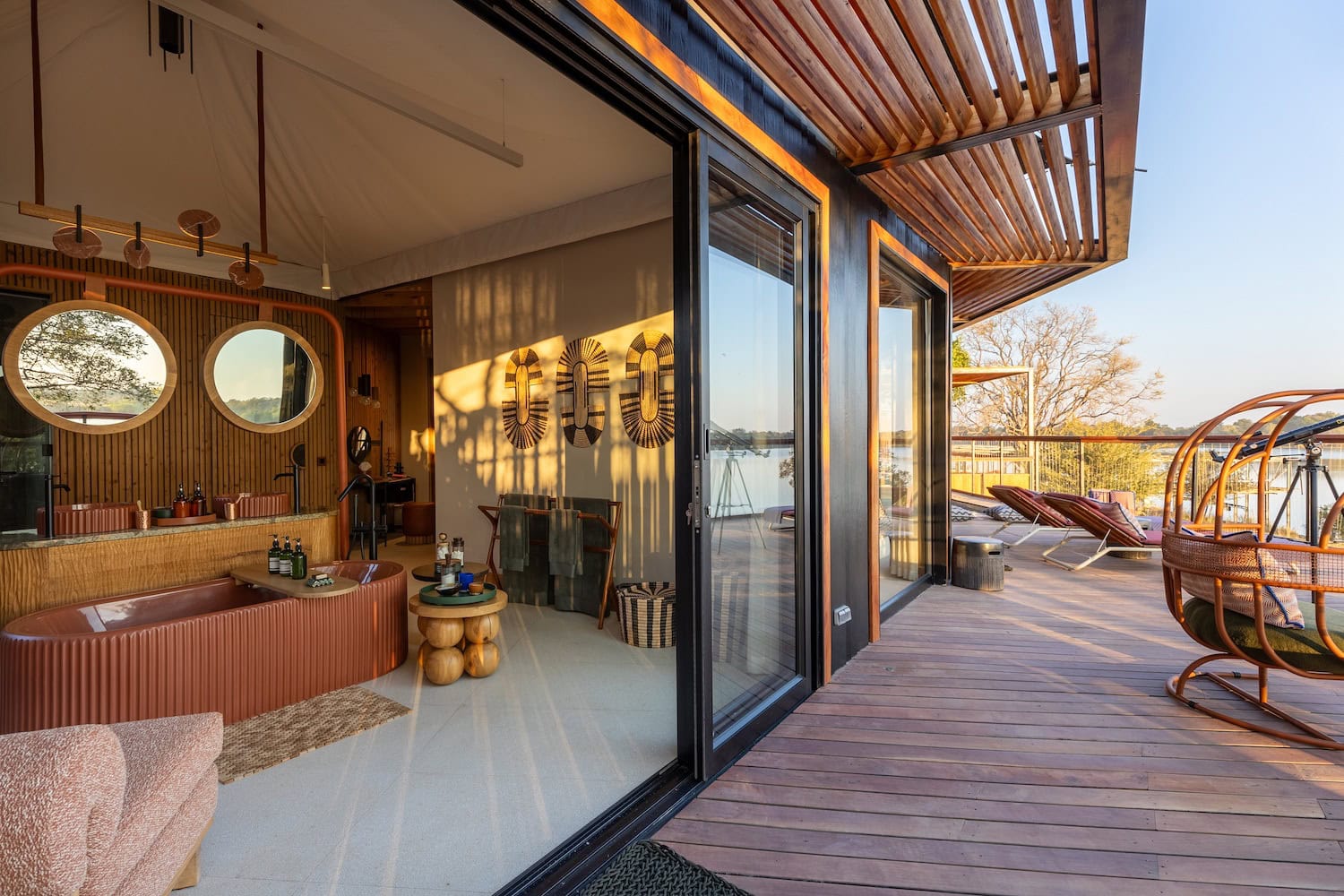Badezimmer mit weit geöffneten Türen zur Terrasse im Anantara Kafue River Camp, im Vordergrund eine braune Badewanne aus welligen Fliesen, auf der Terrasse stehen Möbel, die auf einen Seeblick ausgerichtet sind.