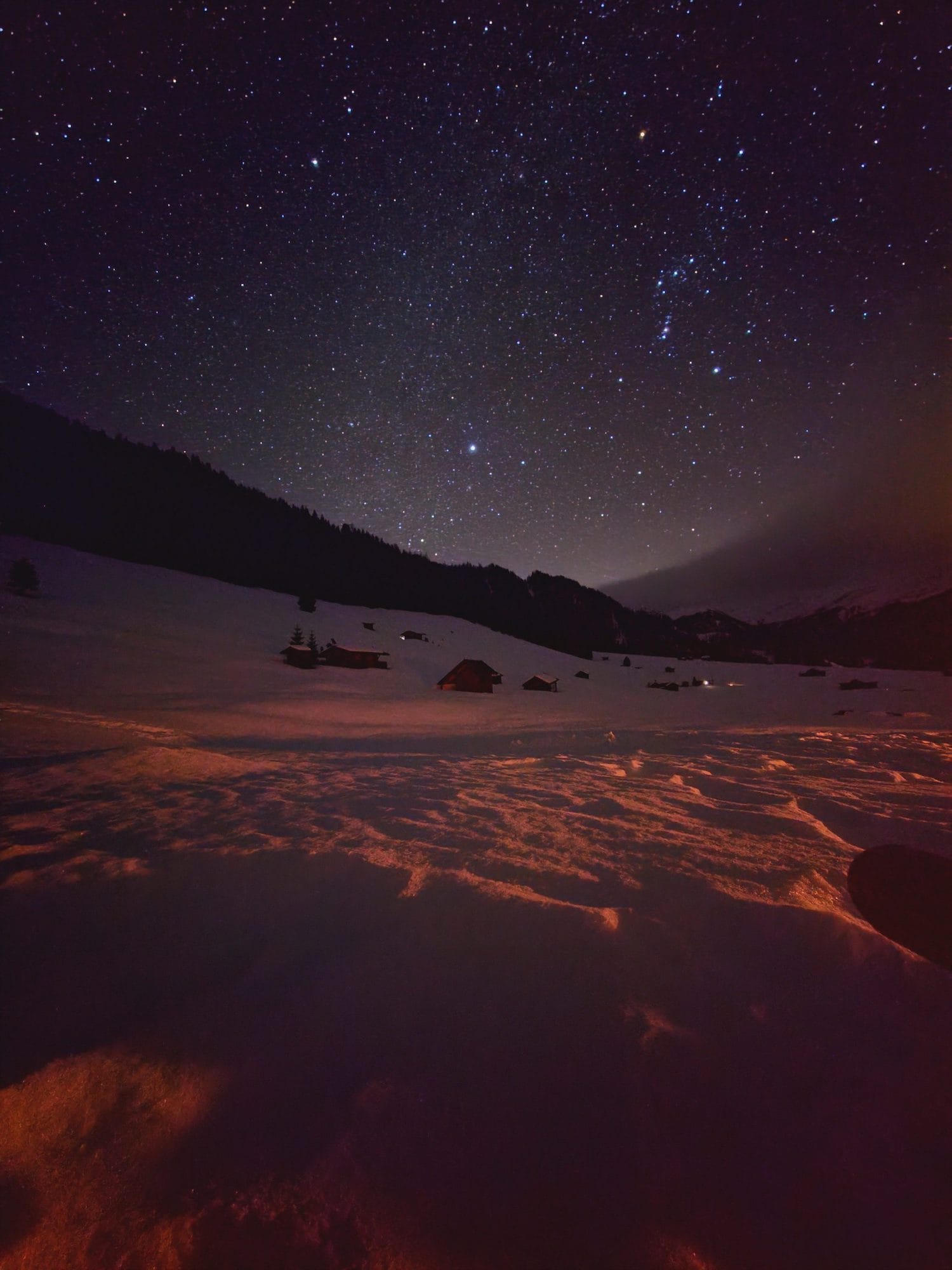 Sternenhimmel über Tirol im Winter, im Vordergrund ein schneebedecktes Feld, im Hintergrund ein Berg, alles ist dunkel, die Sterne funkeln und die Natur ist ein rotes Abendlicht gehüllt.